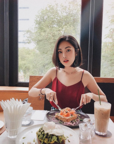 A young woman in a red dress sitting at a restaurant table with a meal of salmon, a drink, and what appears to be a matcha dish, with greenery visible through the window behind her.
