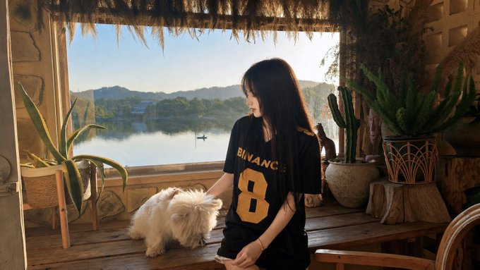 The image features a woman standing near a window, holding her dog outside. The scene takes place next to a lake or river, with a boat visible in the water. There are also potted plants nearby, adding greenery to the area. The photo is related to the post because it appears that the woman and her dog have been featured on this image.
