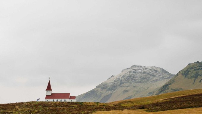 A white church with a red roof and steeple sits in a grassy landscape with a large mountain in the background under an overcast sky, with a small flag visible near the church.