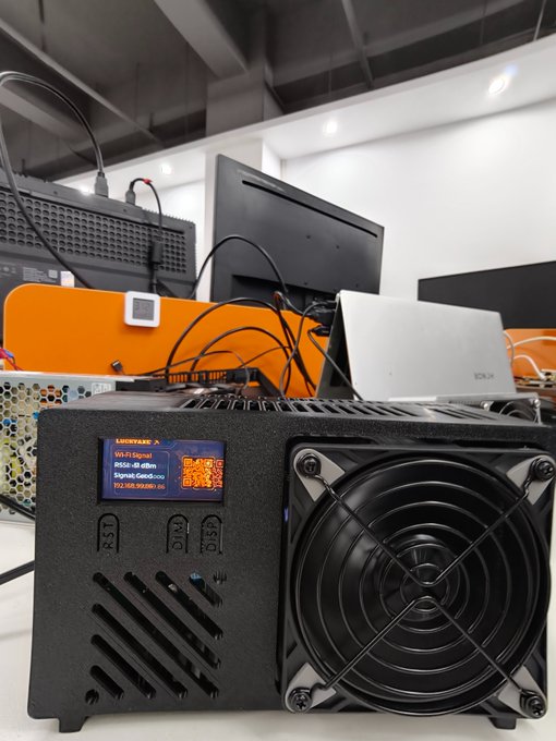 A close-up of a black electronic device, likely a Bitcoin miner, featuring a cooling fan and a small LCD screen displaying 'LUCKYSTAR', 'Wi-Fi Signal', 'IP5528-49.abhm', and 'Signal: Good'.