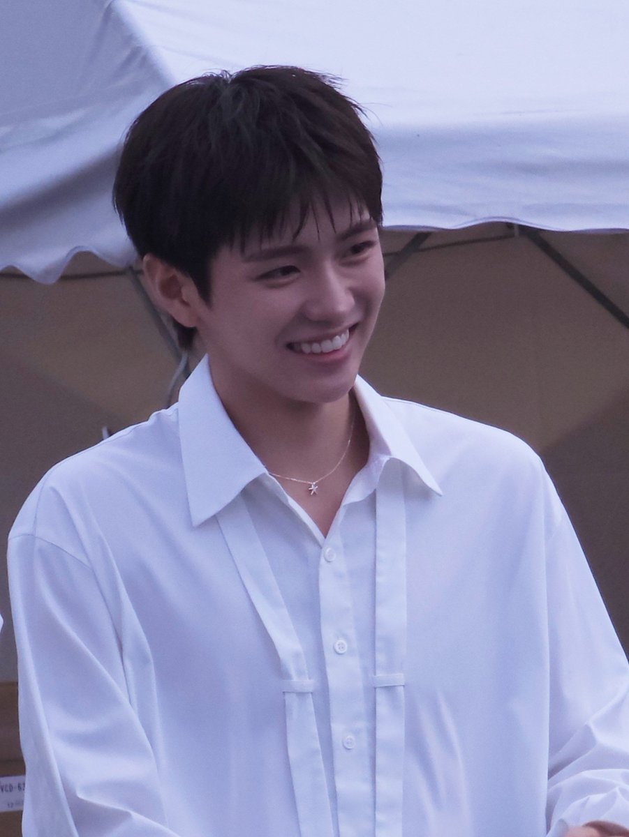 A smiling young man with dark hair wearing a white button-up shirt and a delicate cross necklace, photographed against a light-colored background.