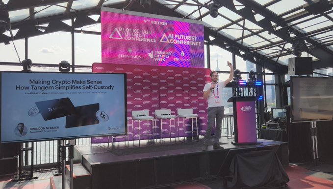 A presenter from Tangem stands on stage at the Blockchain Futurist Conference, displaying a slide titled "Making Crypto Make Sense: How Tangem Simplifies Self-Custody," illustrating the company's presence at the event. Several white chairs are set up on stage next to a podium branded with the conference logo, while attendees in the crowd appear engaged with the presentation.