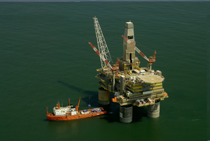 An aerial view of an offshore oil rig in green water with cranes, a helipad, and an orange supply vessel docked alongside it.