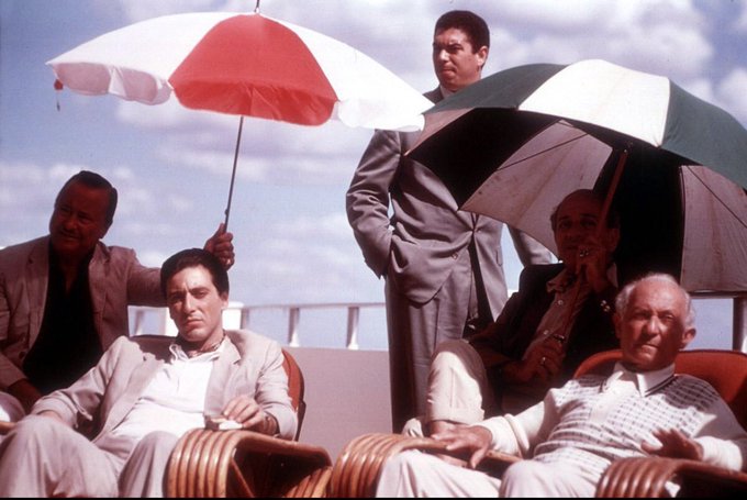 A group of well-dressed men lounge in deck chairs under red, white, and green striped umbrellas on a sunny rooftop, evoking a vintage cinematic atmosphere. The luxurious, leisurely setting of the scene mirrors the laid-back yet exclusive vibe of the post, which hints at an upcoming tokenization event with a teaser of "coming soon" tied to the @base community.