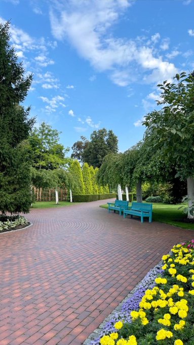 A peaceful park pathway paved with red bricks curves through lush greenery, featuring bright blue benches and vibrant yellow and purple flowers under a partly cloudy blue sky.
