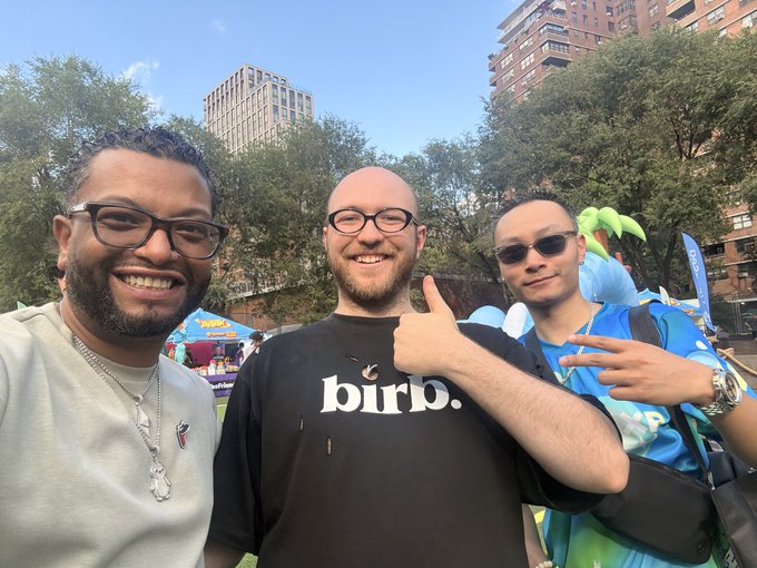 Three men pose together outdoors at what appears to be a music festival in a city park, with the middle man wearing a black 'bird.' t-shirt and giving a thumbs up.