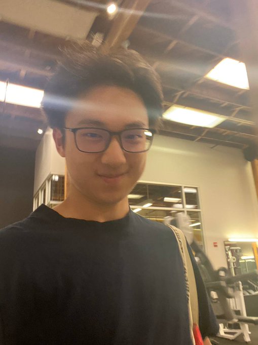 A selfie of a young man with dark hair and glasses wearing a black t-shirt, taken in what appears to be a gym with industrial ceilings and exercise equipment in the background. The caption humorously compliments him as having "CEO energy on the outside, golden retriever on the inside" while jokingly lamenting not having seen his abs yet.