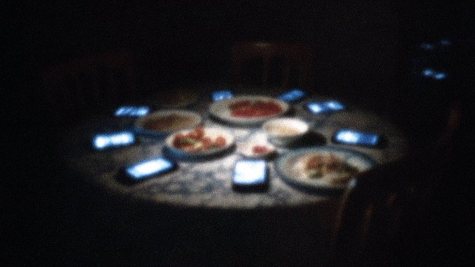 A dimly lit round dining table with plates of food surrounded by multiple smartphones with glowing screens placed face-up where diners would sit.