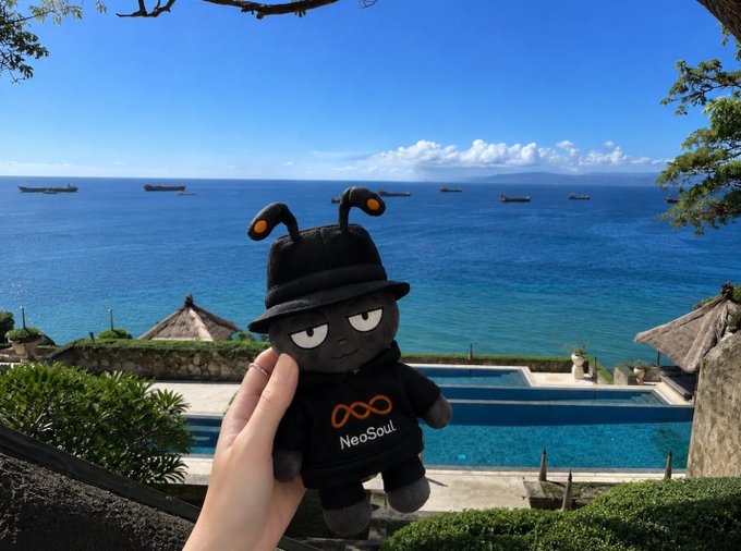 A plush "Antboy" mascot wearing a black hat, branded with "NeoSoul," is held up against a stunning ocean view with ships in the distance and an infinity pool below—capturing the tranquil "weekend reset" moment before the EVOEVO 2.0 launch.