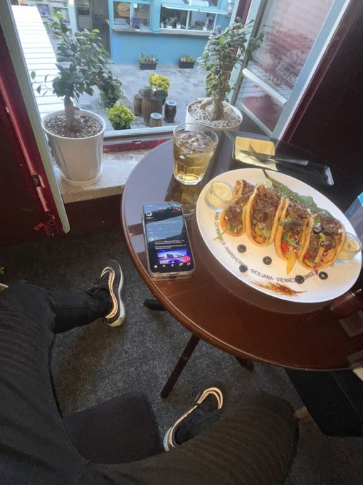 A person sits at a café table with their feet up, enjoying a plate of four loaded beef tacos served with a side of vegetables, alongside a glass of beer and a smartphone on the table. The scene captures a relaxed Tuesday lunch, with potted plants visible through the window and the festive taco spread perfectly matching the "Happy Tuesday 🌮" post.