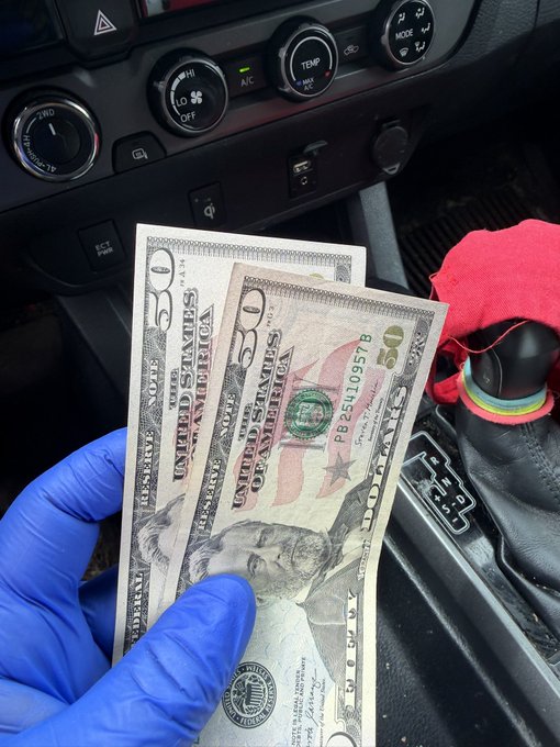 The image shows a person holding two twenty dollar bills in their gloved hand while sitting inside a car. The person appears to be tipping someone, possibly a house cleaner or a service provider, for their work. The car's interior features various controls such as a steering wheel and a gear shift, indicating that the vehicle is parked indoors.
