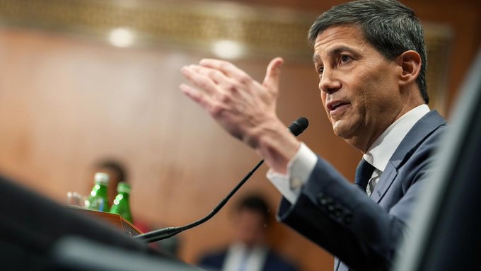 A man in a dark suit gestures with his hand while speaking at a podium with a microphone, appearing to be at a legislative or official hearing.