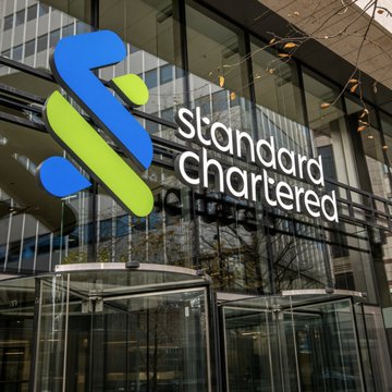The image shows the exterior of a Standard Chartered building featuring the company's blue and green logo prominently displayed alongside its name in white text. The image relates to the post by visually representing Standard Chartered, the banking institution backing Anchorpoint's HKDAP stablecoin initiative for institutional users in Hong Kong.