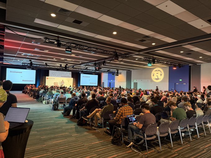 A large audience sits in rows of chairs in a conference hall, facing presentation screens displaying 'Development' and 'Maintenance' slides, with a Rust programming language logo projected on the wall.