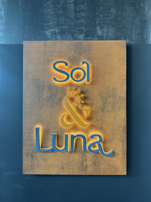 Illuminated sign reading 'Sol' and 'Luna' with a corn plant logo between the words, mounted on a tan panel against a dark background.