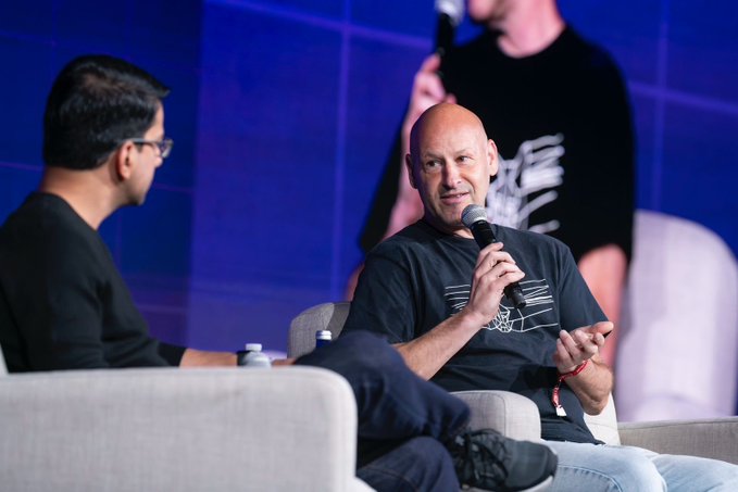 The image shows a man sitting in a chair, holding a microphone, while another person stands behind him. The scene appears to be taking place during an event or conference where speakers are addressing an audience. The main speaker is wearing a black shirt with white text on it and is talking about Ethereum's history and its future. There are other people present in the image as well, suggesting that this might be a larger gathering of attendees at the event.