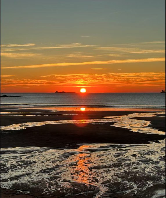 A stunning sunset over the ocean, with the sun reflecting off the wet sand and shallow waters of a beach under a colorful sky.