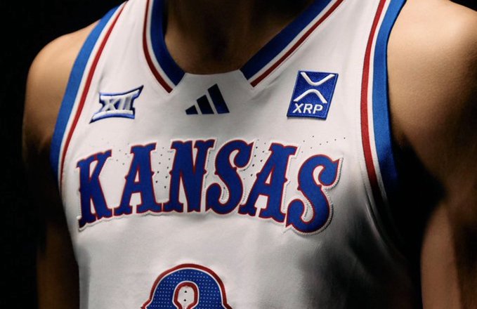 A close-up of a Kansas University basketball jersey featuring the "XRP" logo patch alongside the Big 12 conference emblem and Adidas logo, with "KANSAS" prominently displayed in blue lettering. The image visually represents the historic partnership where XRP became the first cryptocurrency to be featured on collegiate athletic jerseys, as highlighted in the accompanying post.