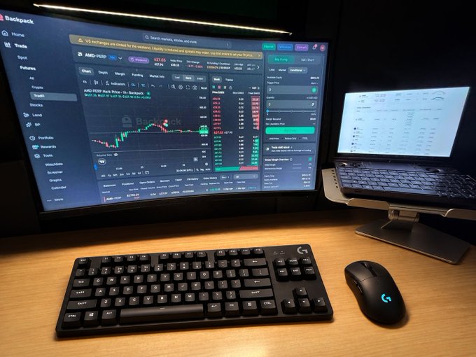 A trading workstation setup featuring a large monitor displaying the Backpack exchange with AMD-PERP showing $37.25 on a candlestick chart, paired with a laptop, Logitech keyboard, and mouse on a wooden desk.