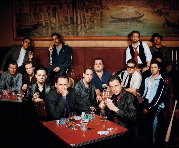 A group photo of approximately twelve men gathered around a red table at what appears to be a poker gathering, with a Venetian-style mural of gondolas in the background.