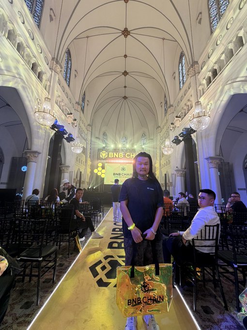 A man with long hair stands on a golden runway at what appears to be a BNB Chain event held inside a stunning cathedral-like venue with soaring vaulted ceilings and ornate chandeliers, holding a branded BNB Chain gift bag while the @pearbinance community event takes place around him.