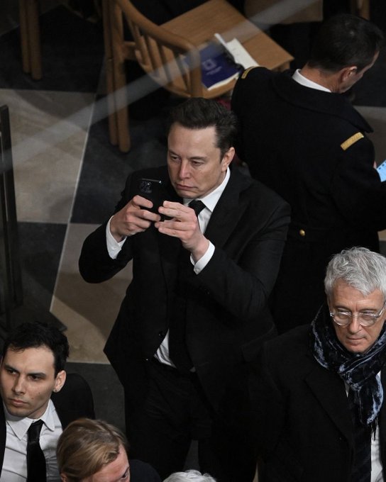A photo of Elon Musk in a black suit and tie looking down at his smartphone, with another person in uniform visible behind him and an older gentleman in glasses to his right.