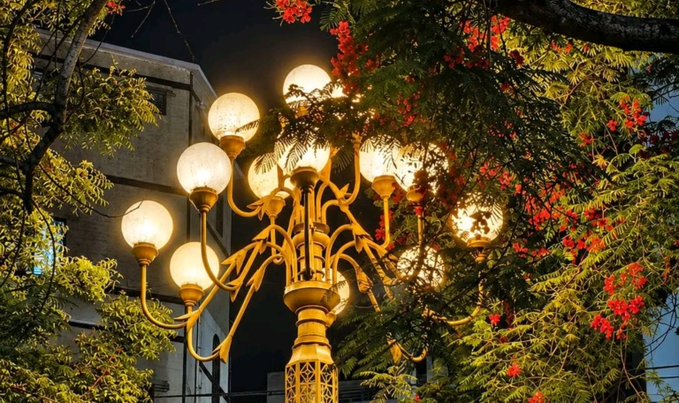 An ornate golden street lamp with multiple glowing globe lights is surrounded by tree branches bearing bright red flowers against a twilight sky.