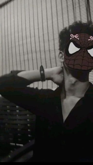 A black-and-white selfie of a person posing with their hands behind their head, wearing a digitally applied Spider-Man mask with cute pink bow decorations.