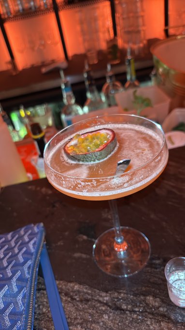 A pink cocktail in a coupe glass garnished with a passion fruit slice, served at a bar with warm orange lighting and bottles visible on shelves in the background.