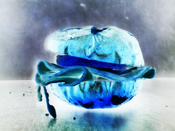 A close-up, cool-toned photograph of a blue and white macaron filled with dark blue cream, with one half melting or dripping downwards onto a reflective surface.