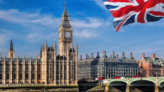 A scenic view of the Palace of Westminster and Big Ben in London, with the British Union Jack flag waving prominently in the foreground.