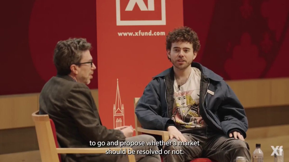 The image shows two men sitting across from each other at a table with chairs. One man has curly hair wearing glasses and is dressed in a suit, while the other man has short hair and is wearing a t-shirt. They are engaged in conversation, possibly discussing a topic related to the post they just saw on social media. The image also contains text that reads "to go and propose whether a market should be resolved or not".