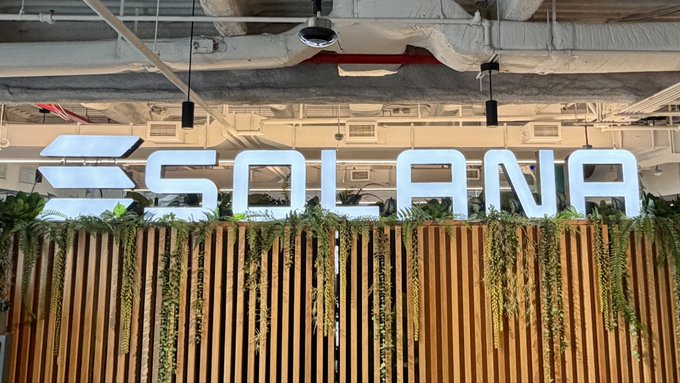 An indoor photo of a large illuminated 'SOLANA' sign with its logo, mounted on a wooden slat wall with hanging green plants and exposed pipes above.