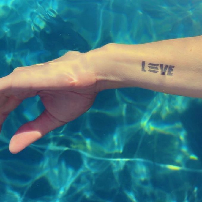 The image features a person's arm with a tattoo of the word "love" written in black ink, located near their wrist. The background of the photo shows water, possibly an ocean or a swimming pool, which adds a sense of tranquility to the scene. The caption provided after the image reads, "realizing I’m on my own eat pray love arc this summer eat in Italy pray with my dao in Amsterdam soon: love in Berlin @SuperteamDE Solana Summit Germany see you there?".
