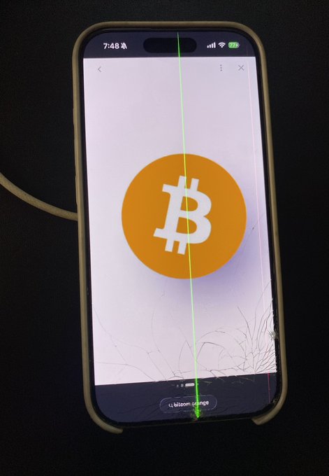 A smartphone displaying a cracked screen showing the orange Bitcoin logo, with the time 7:48 visible at the top.