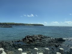A scenic coastal view showing a rocky shoreline with dark stones in the foreground, turquoise water, and a green headland stretching into the distance under a blue sky.