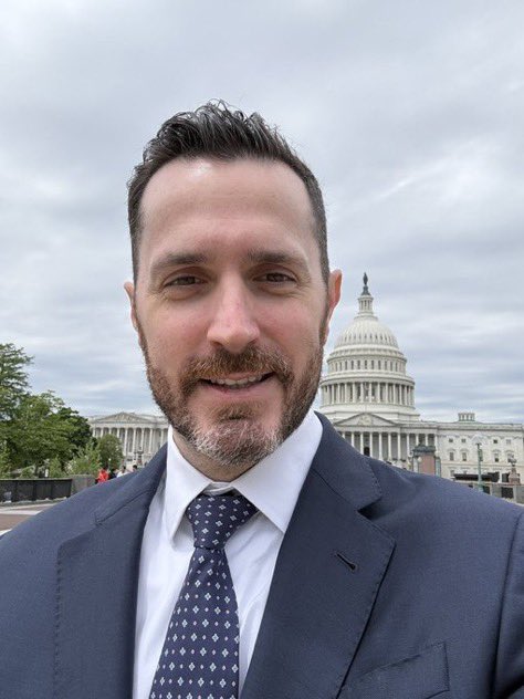 A selfie of a man in a suit and tie smiling in front of the U.S. Capitol building.
