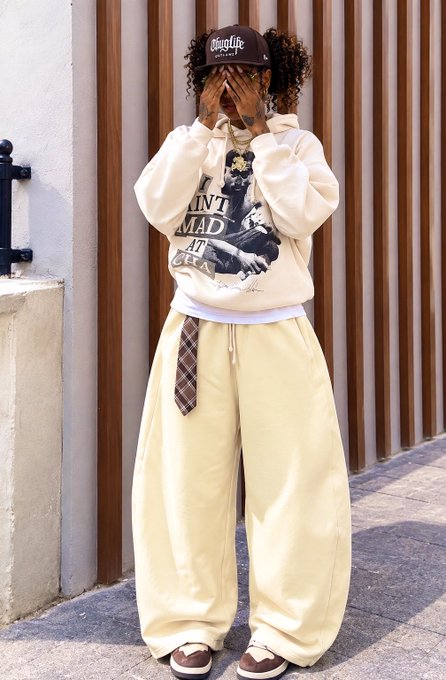 A person models a streetwear outfit consisting of a cream hoodie with a graphic, yellow baggy pants, a checkered tie, and a brown cap, standing against a brown slatted wall.
