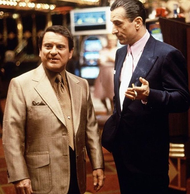Two men in suits stand together in a casino setting, one holding a cigarette.