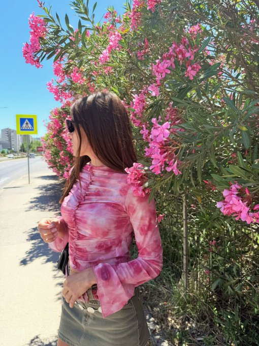 The image features a woman wearing a pink top with a tie-dye pattern, standing next to a tree filled with pink flowers. The background of the photo includes a street sign and buildings, suggesting that she might be in an urban setting. The caption provided seems unrelated to the main subject matter but is likely meant to provide context or additional information about the image.