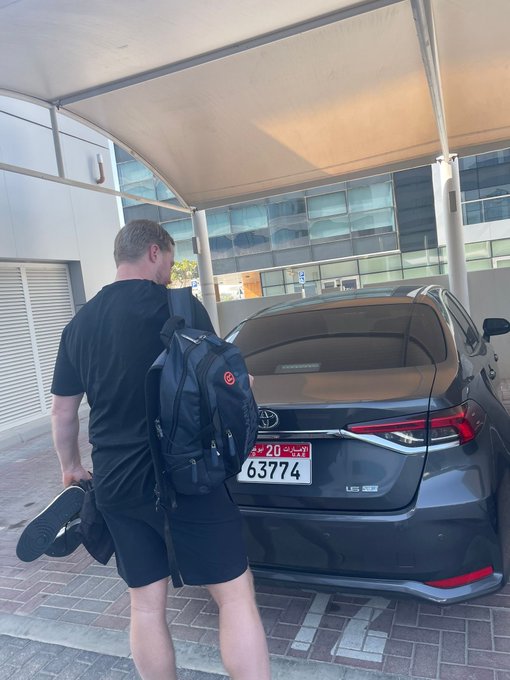 A man wearing a black shirt, shorts, and backpack stands next to a gray Toyota sedan with a Saudi Arabia license plate (63774), holding a pair of shoes under a covered walkway.