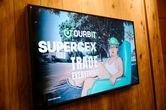 A wall-mounted TV displaying an advertisement for 'OURBIT SUPERCEX - TRADE EVERYTHING' featuring a man sitting in a green chair.