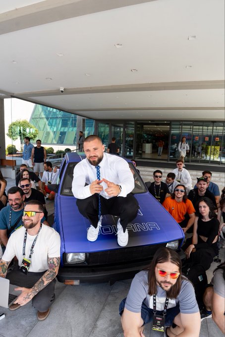 Group photo of people gathered around a blue car at an outdoor event, with a man in a white shirt and tie sitting on the hood.