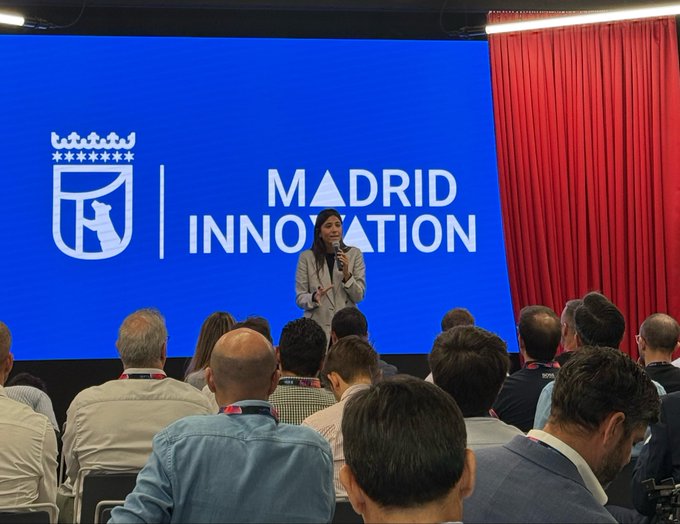 A woman speaks at a podium in front of a large blue screen displaying 'MADRID INNOVATION' alongside a coat of arms, with red curtains flanking the stage and an audience seated before her.