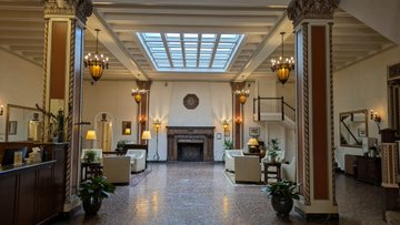 An elegant hotel lobby interior with classical architecture, featuring tall columns, a coffered skylight, a fireplace at the far end, and arranged lounge seating.