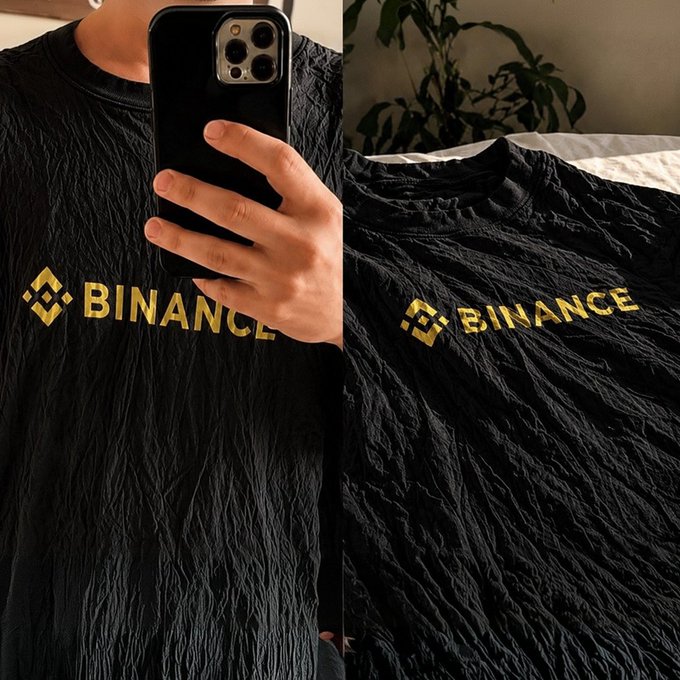 A side-by-side mirror selfie and close-up showing a black wrinkled t-shirt featuring the yellow "BINANCE" logo and crypto exchange emblem, humorously fitting the caption's reference to wrinkled tees trending as fashion.