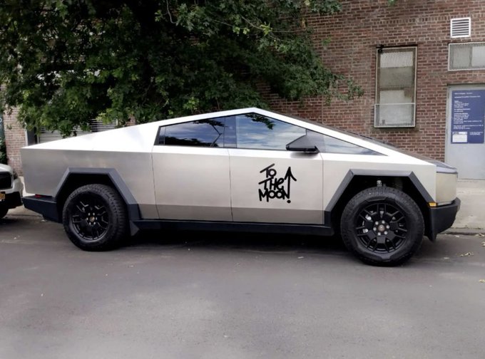 The image features an older model truck parked on the side of the street with graffiti on its door. The truck has a "To a man" sticker on the door, which adds a personal touch to the vehicle. The truck appears to be silver in color, and there is a tree nearby that provides some greenery to the scene.