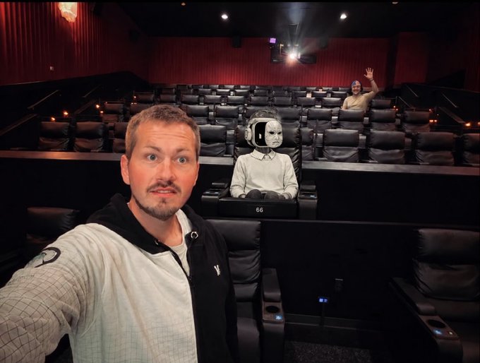 A man takes a selfie in a mostly empty movie theater, with an AI-generated or superimposed image of a child wearing a VR headset sitting in the seat behind him and another person waving in the back row.