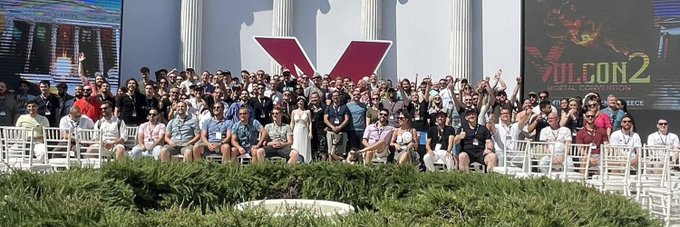 A large group photo of attendees seated and standing on outdoor bleachers at what appears to be the Vulcan 2 production kickoff event, with a large red 'V' logo visible on the backdrop.