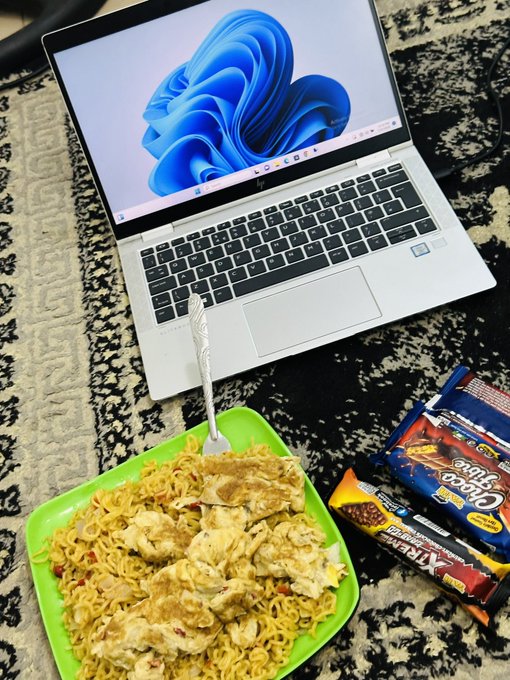 A laptop displaying a blue Windows wallpaper sits on a carpet next to a green plate of instant noodles with a fork, alongside Choco Pie and Xingyu snack packages.